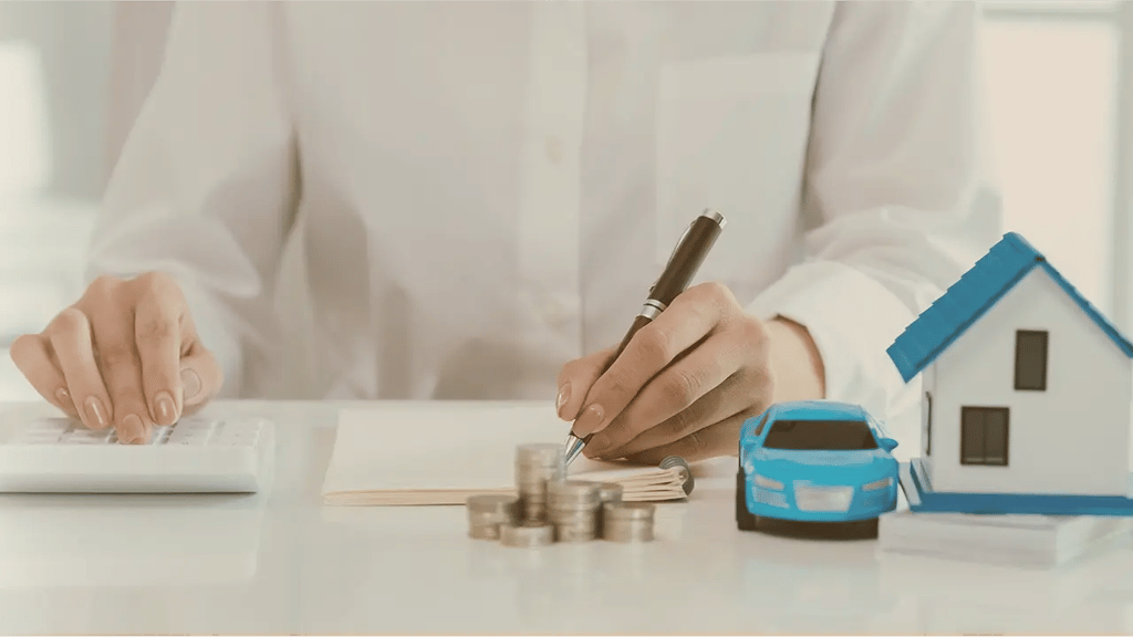 Persona calculando finanzas personales junto a una casa y un coche en miniatura, representando los seguros de ahorro más rentables.