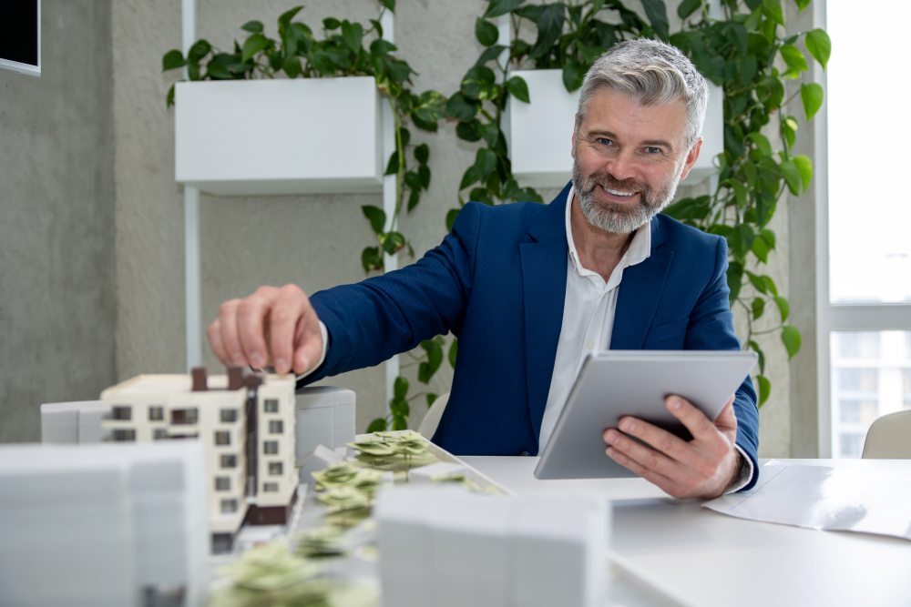 Administrador de fincas profesional sonriendo en una oficina luminosa mientras revisa la gestión de propiedades en una tablet junto a la maqueta de un edificio