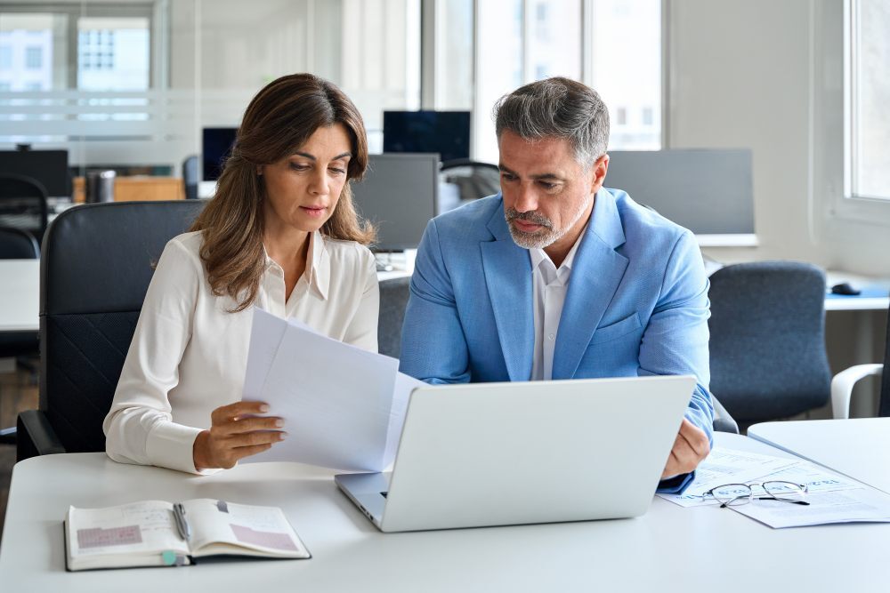 Dos profesionales con perfil directivo analizando y comparando detalladamente contratos de seguros sobre un escritorio en una oficina luminosa.