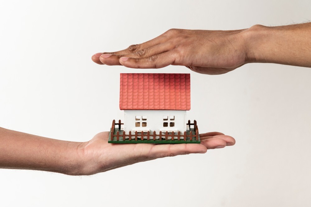 Dos pares de manos protegiendo y sosteniendo una casa en miniatura para representar las coberturas de un seguro de hogar para comunidades.