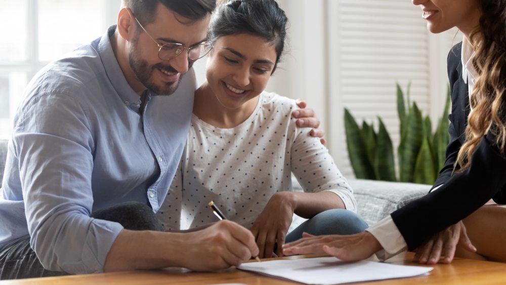 Pareja feliz firmando un contrato de protección para su vivienda asesorados por un agente profesional en un entorno luminoso.